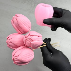 Pu&ograve; includere: Stampi rosa a forma di tulipano e fiori finiti. I fiori sono su spiedini di legno. Gli stampi sono tenuti da una persona con guanti neri. Lo sfondo &egrave; grigio chiaro.