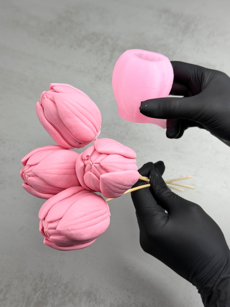 Pu&ograve; includere: Stampi rosa a forma di tulipano e fiori finiti. I fiori sono su spiedini di legno. Gli stampi sono tenuti da una persona con guanti neri. Lo sfondo &egrave; grigio chiaro.