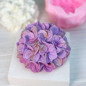 May include: A detailed, purple and pink hydrangea flower sculpture sits on a white square base. The flower has multiple layers of petals with subtle color variations. In the background, other floral sculptures in white and pink are slightly out of focus.
