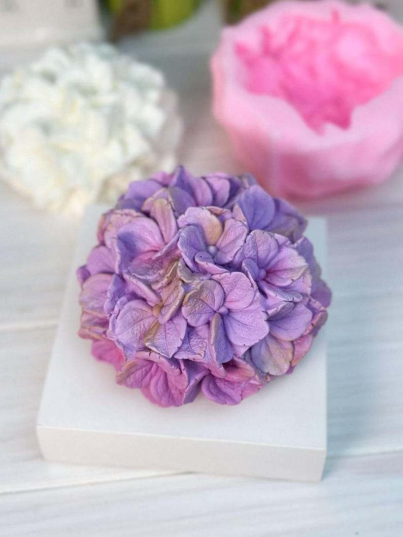 May include: A detailed, purple and pink hydrangea flower sculpture sits on a white square base. The flower has multiple layers of petals with subtle color variations. In the background, other floral sculptures in white and pink are slightly out of focus.