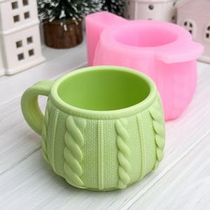 Könnte beinhalten: Eine hellgrüne Tasse mit Zopfmuster und Henkel. Dahinter befindet sich eine rosa Tassenform. Die Tasse steht auf einer weißen Holzoberfläche. Miniaturhäuser und ein kleiner Weihnachtsbaum sind im Hintergrund.