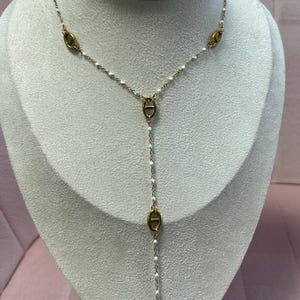 May include: Gold-toned necklace with a Y-shaped design. The necklace features oval-shaped pendants and small white beads. The necklace is displayed on a mannequin with a light pink background.