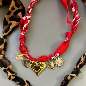以下が含まれることがあります： 生地のチャームが付いた金のネックレスが2本。1本のネックレスには赤いバンダナ生地のチャーム、もう1本にはレオパード柄の生地のチャームが付いています。両方のネックレスには、金のハート、星、月、太陽のチャームが付いています。