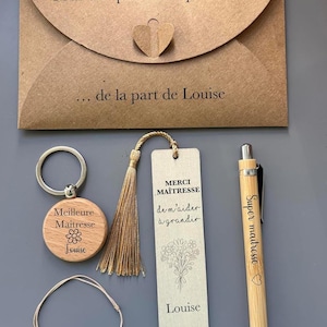 Könnte beinhalten: Ein Geschenkset mit einem braunen Umschlag, einem Holzschlüsselanhänger, einem Lesezeichen, einem Stift und einem Armband. Der Schlüsselanhänger und das Lesezeichen haben Text. Der Stift ist bambusfarben. Das Armband hat weiße Perlen mit dem Wort "MERCI".