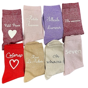 Può includere: Una collezione di otto calzini colorati con texture glitterate. Ogni calzino presenta un testo unico in scrittura bianca, tra cui frasi come "Petit Prince", "Petite Jeanne" e "Cotorep". Alcuni calzini hanno disegni a cuore.