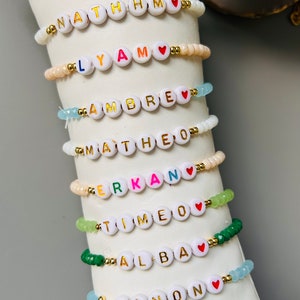 May include: A set of eight colorful beaded bracelets with gold letter beads spelling out names. Each bracelet has a small red heart charm.