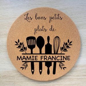 Peut inclure: Un dessous de plat rond en liège avec le texte "Les bons petits plats de Mamie Francine" et une silhouette noire d'ustensiles de cuisine, dont une spatule, une cuillère, un rouleau à pâtisserie et un fouet.