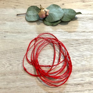 Pode incluir: Um conjunto de pulseiras de cordão vermelho, amarradas juntas em um nó. As pulseiras são feitas de cordão vermelho fino e estão dispostas em círculo.