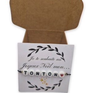 May include: A white card with black text and a black string bracelet with white beads spelling "TONTON" and a red heart bead. The card says "Je te souhaite un Joyeux Noël mon..."