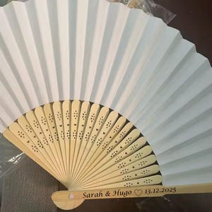 Peut inclure: Éventail en papier blanc avec un manche en bois clair. L'éventail est ouvert, révélant son design plissé. Le manche est gravé des noms "Sarah & Hugo" et de la date "13.12.2025".