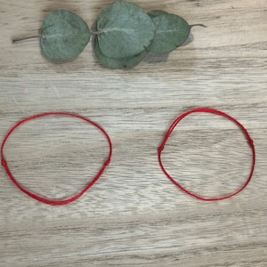 Könnte beinhalten: Zwei rote Armbänder aus Schnur auf einer Holzoberfläche. Die Armbänder sind einfach und aus einem einzigen roten Faden gefertigt.