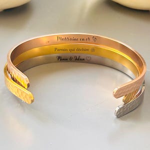 Könnte beinhalten: Drei übereinanderliegende Armbänder aus Gold, Silber und Roségold mit graviertem Text. Das obere Armband trägt die Aufschrift "Marraine en or", das mittlere Armband die Aufschrift "Parrain qui dechire" und das untere Armband die Aufschrift "Manon & Julian".