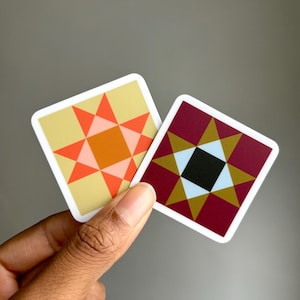 May include: Two square stickers with geometric star designs. One sticker has a yellow background with orange and coral accents. The other has a burgundy background with gold and black accents. Both have white borders.