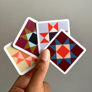 May include: A set of four square stickers featuring geometric star patterns in various color combinations including maroon, light blue, orange, red, and cream. Each sticker has a white border and displays a distinct, symmetrical quilt-inspired design.