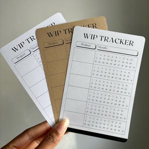 May include: Three work-in-progress (WIP) tracker cards in white and kraft paper. Each card has the text "WIP TRACKER", with sections for "Project" and "Month", and a calendar grid. The cards are held in a hand.