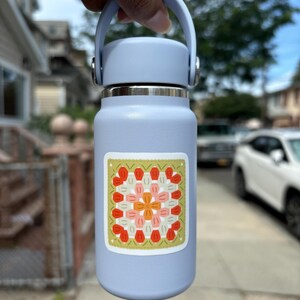 May include: A pale blue insulated water bottle with a matching handle and lid. A square sticker with a floral design in orange, pink, and red is affixed to the bottle. The bottle is approximately 25 cm tall.
