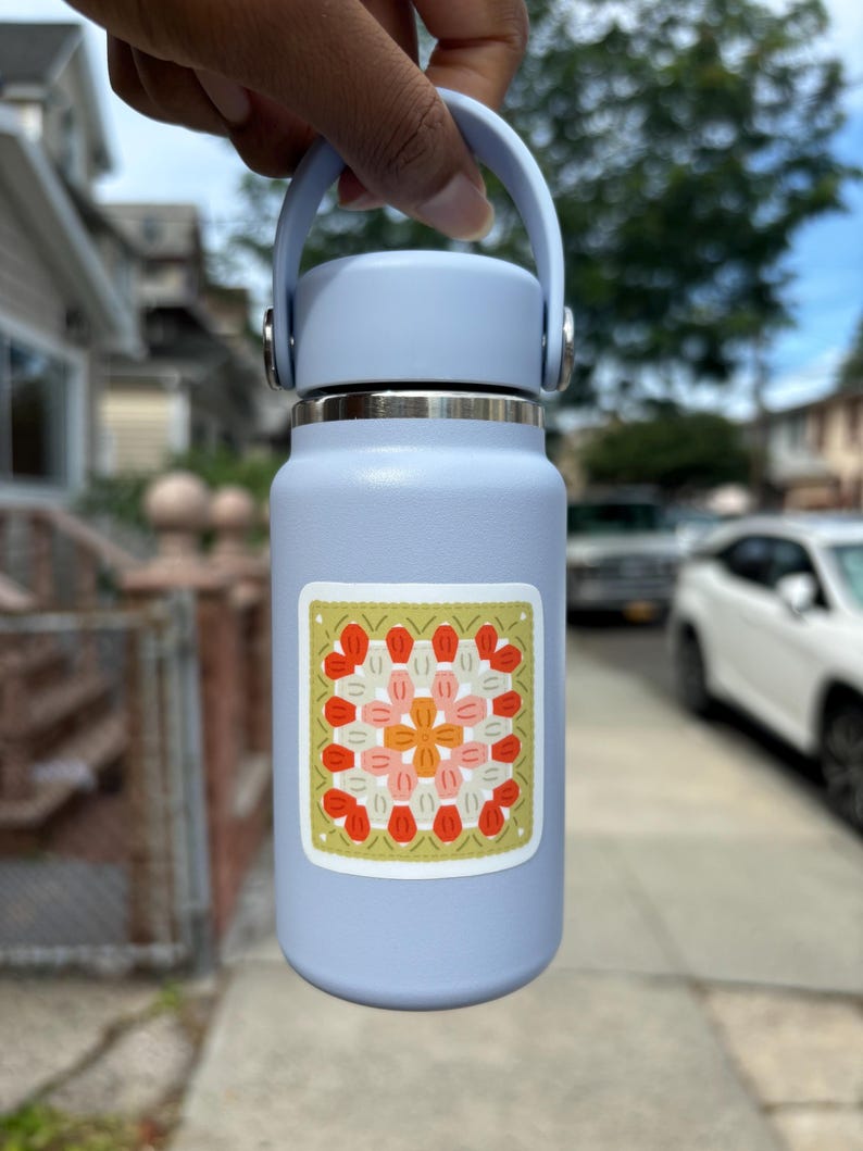 May include: A pale blue insulated water bottle with a matching handle and lid. A square sticker with a floral design in orange, pink, and red is affixed to the bottle. The bottle is approximately 25 cm tall.