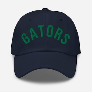 May include: A navy blue baseball cap with the word "GATORS" embroidered in green across the front. The cap has a curved brim and a fabric-covered adjustable strap.