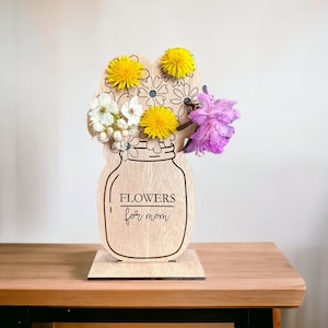 May include: Wooden flower vase with the text "Flowers for Mom" engraved on the front. The vase is decorated with a cutout design of a mason jar and flowers. The vase is standing on a wooden base.