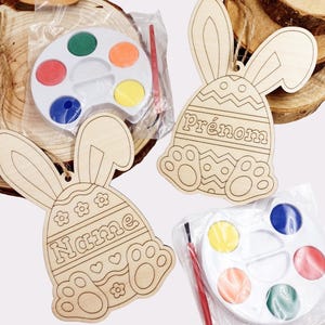 Può includere: Due decorazioni per uova di Pasqua a forma di coniglio in legno con palette di colori e pennelli. Le uova di coniglio hanno le parole "Name" e "Prénom" su di esse.