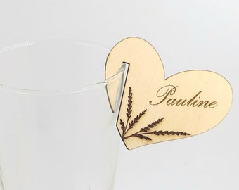 May include: Wooden heart-shaped place card with a wheat stalk design and the name "Pauline" engraved on it. The place card is attached to a clear glass.