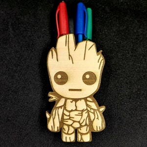May include: Wooden pencil holder shaped like a cartoon character with a green, blue, and red pen sticking out of the top. The character has a brown, textured surface and a neutral facial expression.