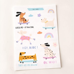 Könnte beinhalten: Ein weißer Notizblock mit Illustrationen von Hunden auf Skateboards. Die Zeichnungen enthalten Sätze wie "Catch Me - If You Can" und "Hey, Dude!" Das Design umfasst Regenbogen, Blumen und die Worte "OK Wau!"
