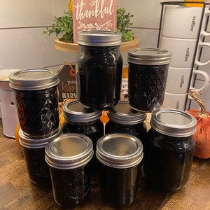 Könnte beinhalten: Eine Sammlung von acht Glasbehältern, gefüllt mit einer dunklen Flüssigkeit, wahrscheinlich hausgemachte Marmelade oder Konserven. Die Behälter sind pyramidenförmig auf einer Holzoberfläche angeordnet. Die Behälter haben silberne Deckel und ein Diamantenmuster auf dem Glas.