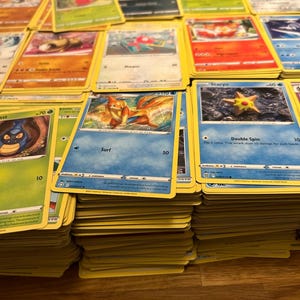 May include: A selection of Pok&eacute;mon trading cards, showcasing diverse characters and artwork. The cards are arranged in stacks and spread out, with yellow borders and vibrant illustrations. Some cards display text such as "Surf" and "Double Spin".
