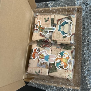 May include: A cardboard box containing four wrapped gifts. Each gift is wrapped in patterned paper and sealed with a clear plastic bag. Stickers of cartoon characters are on the gifts. The box is lined with bubble wrap.