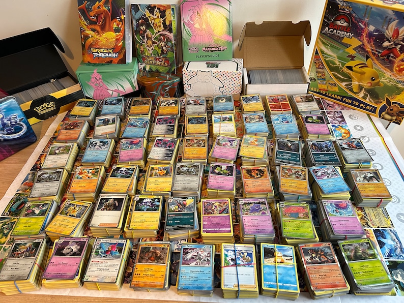 May include: A large collection of Pok&eacute;mon trading cards, organised in stacks and secured with elastic bands. Various card game boxes and accessories are visible, including a "BREAK THROUGH" box and a "Pok&eacute;mon Battle Academy" game.
