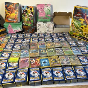 May include: A collection of Pok&eacute;mon trading cards, including stacks of cards, booster packs, and storage boxes. The cards feature various Pok&eacute;mon characters and colourful artwork. A Pok&eacute;mon Academy box is also visible.