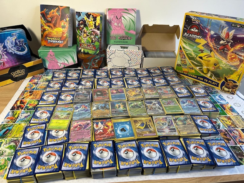 May include: A collection of Pok&eacute;mon trading cards, including stacks of cards, booster packs, and storage boxes. The cards feature various Pok&eacute;mon characters and colourful artwork. A Pok&eacute;mon Academy box is also visible.