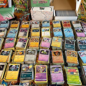 May include: A collection of Pok&eacute;mon trading cards, bound with elastic bands, is shown. Card packs and a player's guide are in the background. The cards display colourful illustrations and text, featuring various Pok&eacute;mon characters.