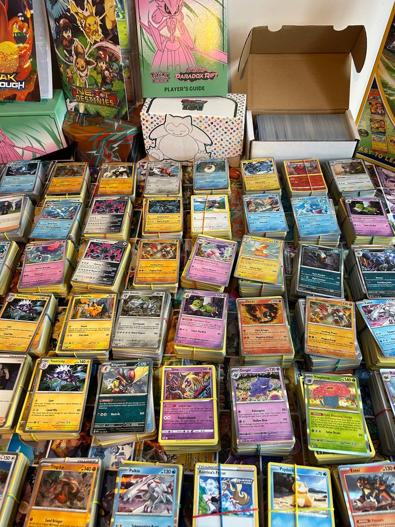 May include: A collection of Pok&eacute;mon trading cards, bound with elastic bands, is shown. Card packs and a player's guide are in the background. The cards display colourful illustrations and text, featuring various Pok&eacute;mon characters.
