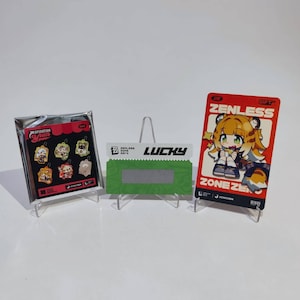 Peut inclure: Trois articles exposés : un paquet scellé de breloques de personnages, une carte verte et blanche "LUCKY", et une carte rouge avec un personnage de dessin animé et le texte "ZENLESS ZONE ZERO". Tous les articles sont sur des présentoirs transparents.