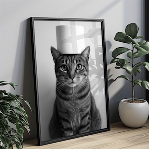 May include: Black and white framed print featuring a tabby cat with a roll of toilet paper balanced on its head. The cat is centered in the frame, with a neutral background. The print is displayed on a wooden surface, with a potted plant visible.