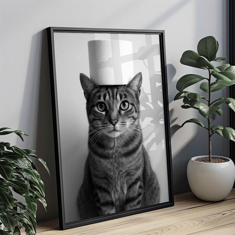 May include: Black and white framed print featuring a tabby cat with a roll of toilet paper balanced on its head. The cat is centered in the frame, with a neutral background. The print is displayed on a wooden surface, with a potted plant visible.