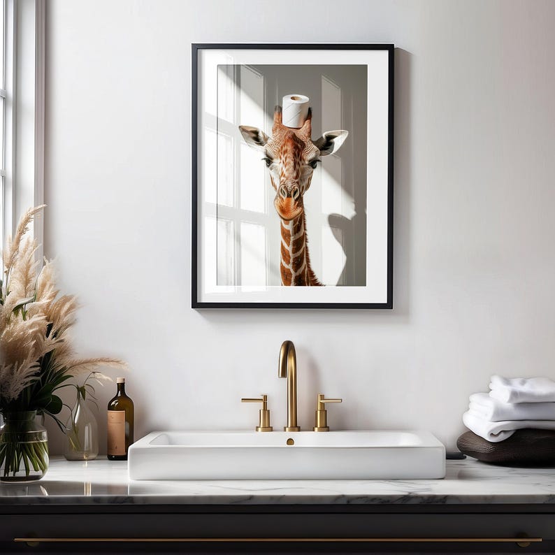 May include: A framed print of a giraffe wearing a toilet paper roll as a hat. The giraffe is looking directly at the camera. The print is hanging on a white wall above a white bathroom sink with gold faucets.
