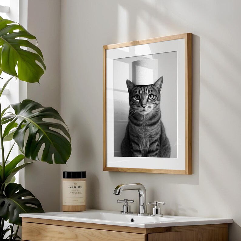 May include: Black and white framed print of a tabby cat with a roll of toilet paper on its head. The print is in a light wood frame and hangs above a bathroom sink. A large green plant is to the left of the print.