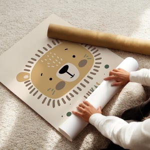 May include: A beige poster with a cartoon illustration of a lion's face. The lion has a brown nose, brown eyes, and a brown mane. The poster is rolled up and being held by a person.