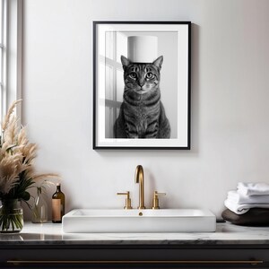 May include: Black and white framed print of a tabby cat with a roll of toilet paper balanced on its head. The cat is centered in the frame, looking directly at the viewer. The print is displayed above a white sink with gold fixtures.