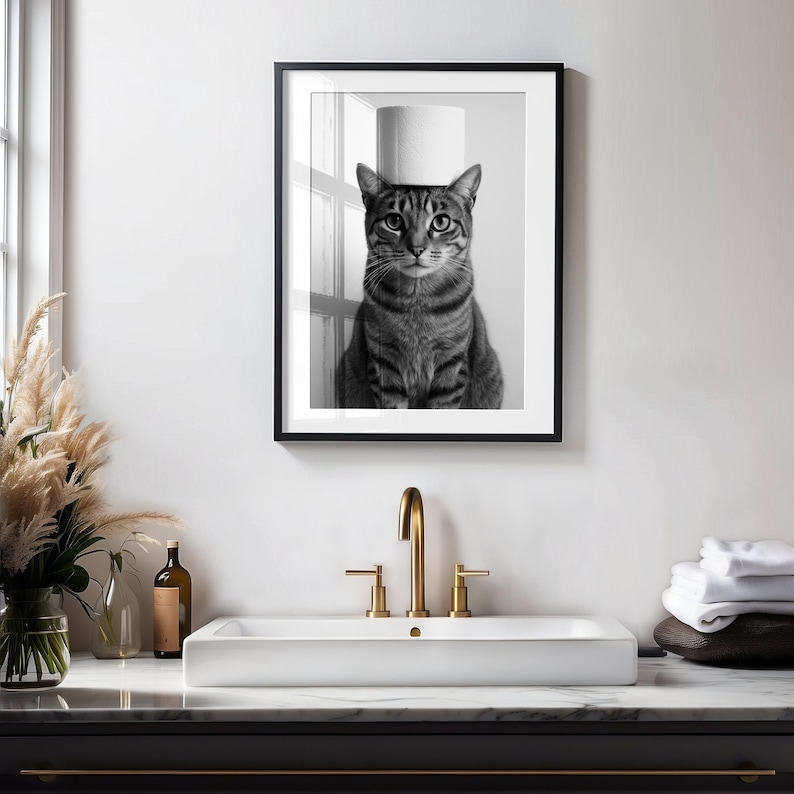 May include: Black and white framed print of a tabby cat with a roll of toilet paper balanced on its head. The cat is centered in the frame, looking directly at the viewer. The print is displayed above a white sink with gold fixtures.