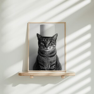 May include: Black and white framed print of a tabby cat with a roll of toilet paper balanced on its head. The cat is centered in the frame, with a neutral background and a wooden shelf below.