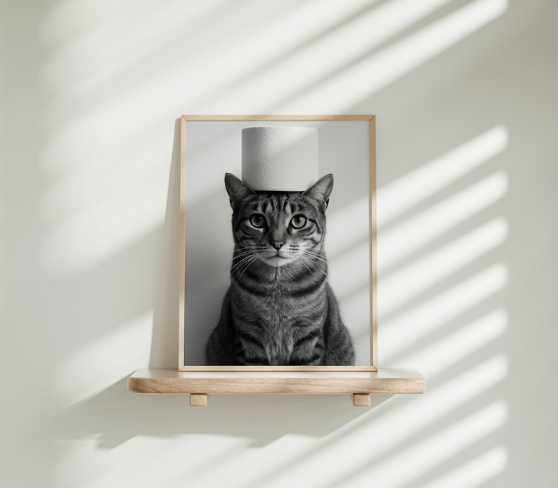 May include: Black and white framed print of a tabby cat with a roll of toilet paper balanced on its head. The cat is centered in the frame, with a neutral background and a wooden shelf below.