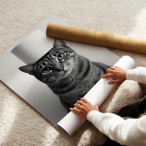 May include: Black and white print of a tabby cat with a curious expression. The print is partially rolled, with a brown paper tube and a white rolled paper tube nearby. The image is on a white surface, and the background is a light-colored carpet.