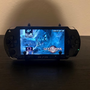 May include: A black Sony PSP handheld gaming console resting on a black stand. The screen displays the God of War: Ghost of Sparta game title, a date of 1/25, and the time 21:48. The device features a directional pad, action buttons, and a screen.