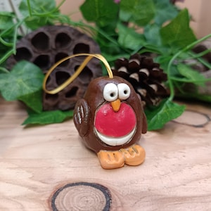 May include: A handmade brown robin ornament with a red breast, white eyes, and orange beak and feet. It has a gold ribbon for hanging. The background features greenery and a pine cone, suggesting a festive theme.