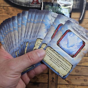 Op de afbeelding: Een hand die een pak fantasie-speelkaarten vasthoudt. De kaarten hebben een blauw-witte hemelachtige achtergrond en bevatten de tekst "Crown of Cyclones" en "Gale". De kaarten liggen op een houten oppervlak.