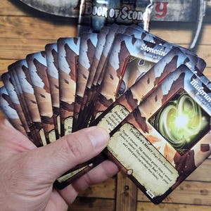 Puede incluir: Una mano sosteniendo un abanico de cartas de juego de fantasía. Las cartas presentan ilustraciones de montañas y orbes brillantes, con texto como "Thulgrun's Mercy" y "Book of Stone". Las cartas tienen un esquema de color marrón y beige.
